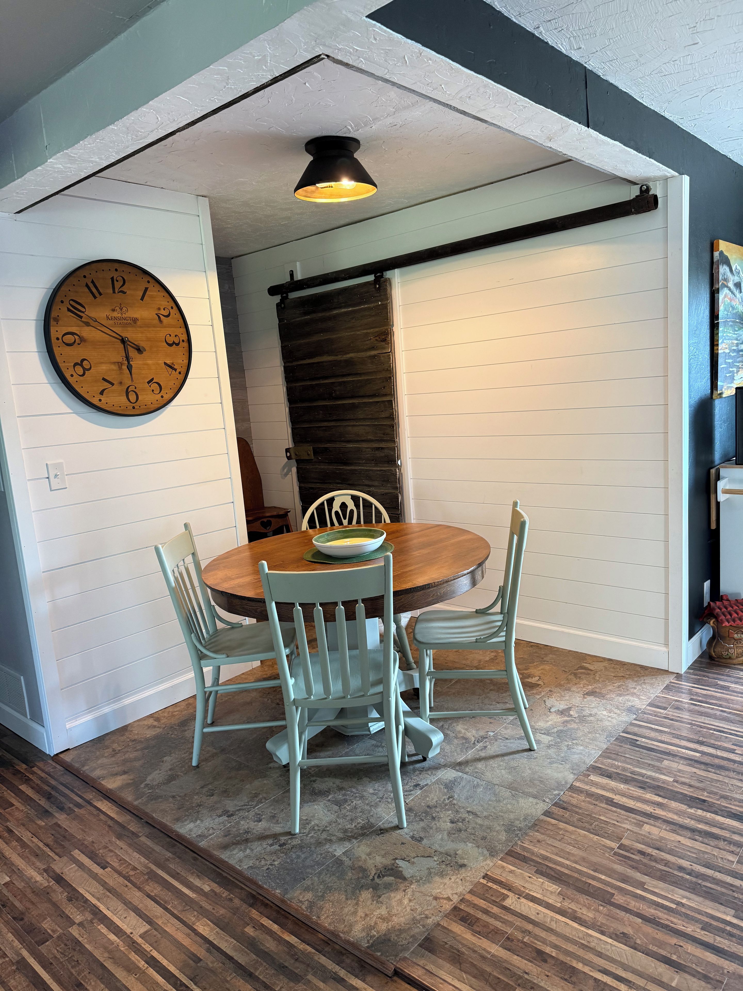 A dining area with a wooden table, four chairs, a wall clock, and a ceiling light fixture.