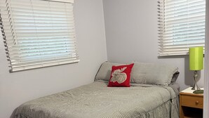 Une chambre à coucher avec un lit, une table de chevet, une lampe et deux fenêtres équipées de stores.