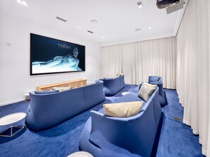 A modern living room with a large flat-screen TV mounted on the wall, a blue sectional sofa, a wooden entertainment unit, and a white curtain. - Tiffany Celeste - Walk to Beach, Water Views, Rooftop Pool (Fort Lauderdale)