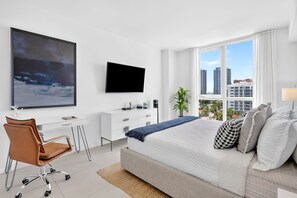 A modern bedroom with a large bed, a desk, a chair, a TV, and a window with a city view. - Tiffany Celeste - Walk to Beach, Water Views, Rooftop Pool (Fort Lauderdale)