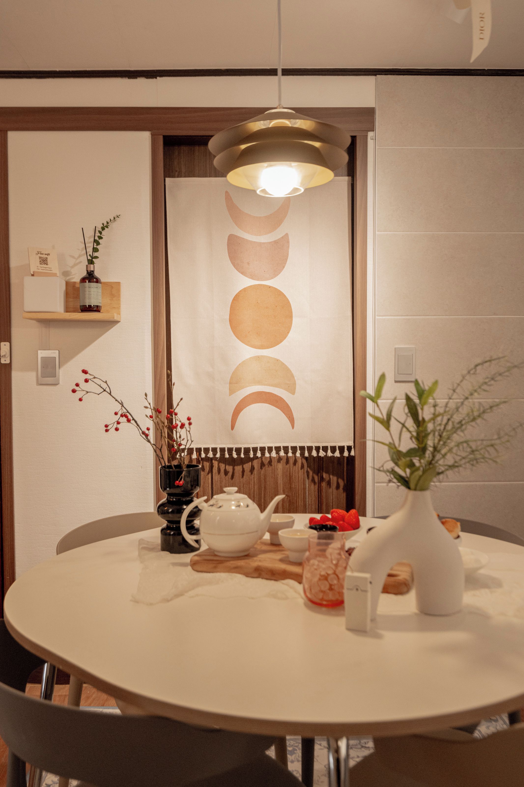 Una mesa redonda para comer con mantel blanco, una tetera negra, una taza de té blanca y un florero blanco con una planta. Hay una estantería en la pared con una botella y una caja pequeña.