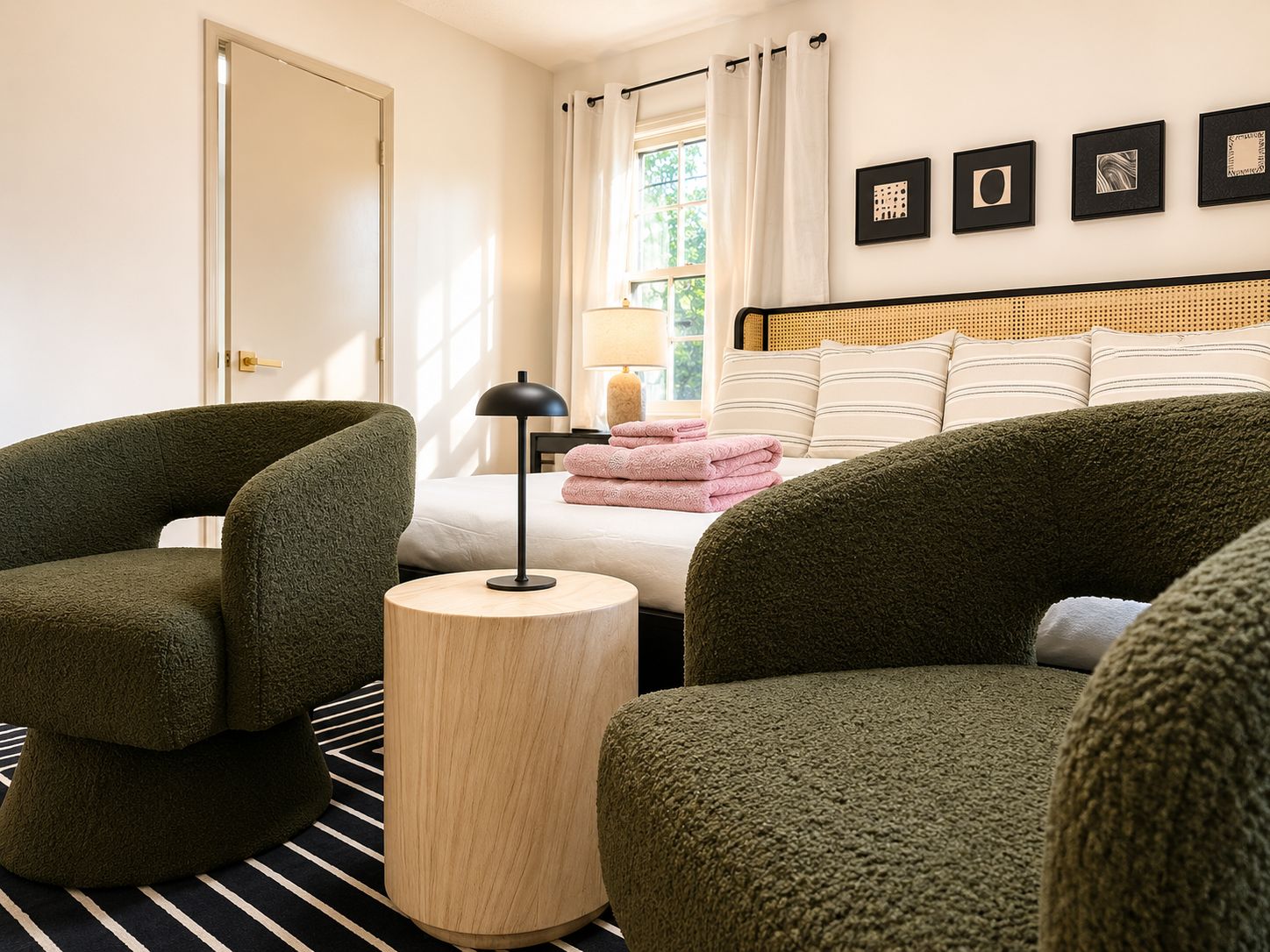 A hotel room with a bed, two green armchairs, a round wooden side table, and a window with curtains.