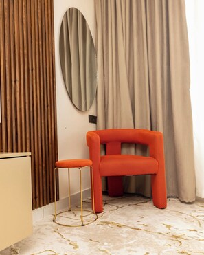 Quarto com parede de painel de madeira, espelho redondo, poltrona vermelha estofada, um pequeno banquinho e chão com padrão marmorizado.