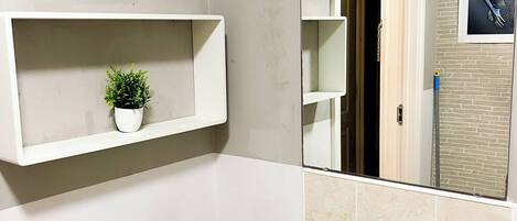 Un baño con un lavamanos blanco, dos estantes blancos con plantas, un espejo y una puerta que da a otra habitación.