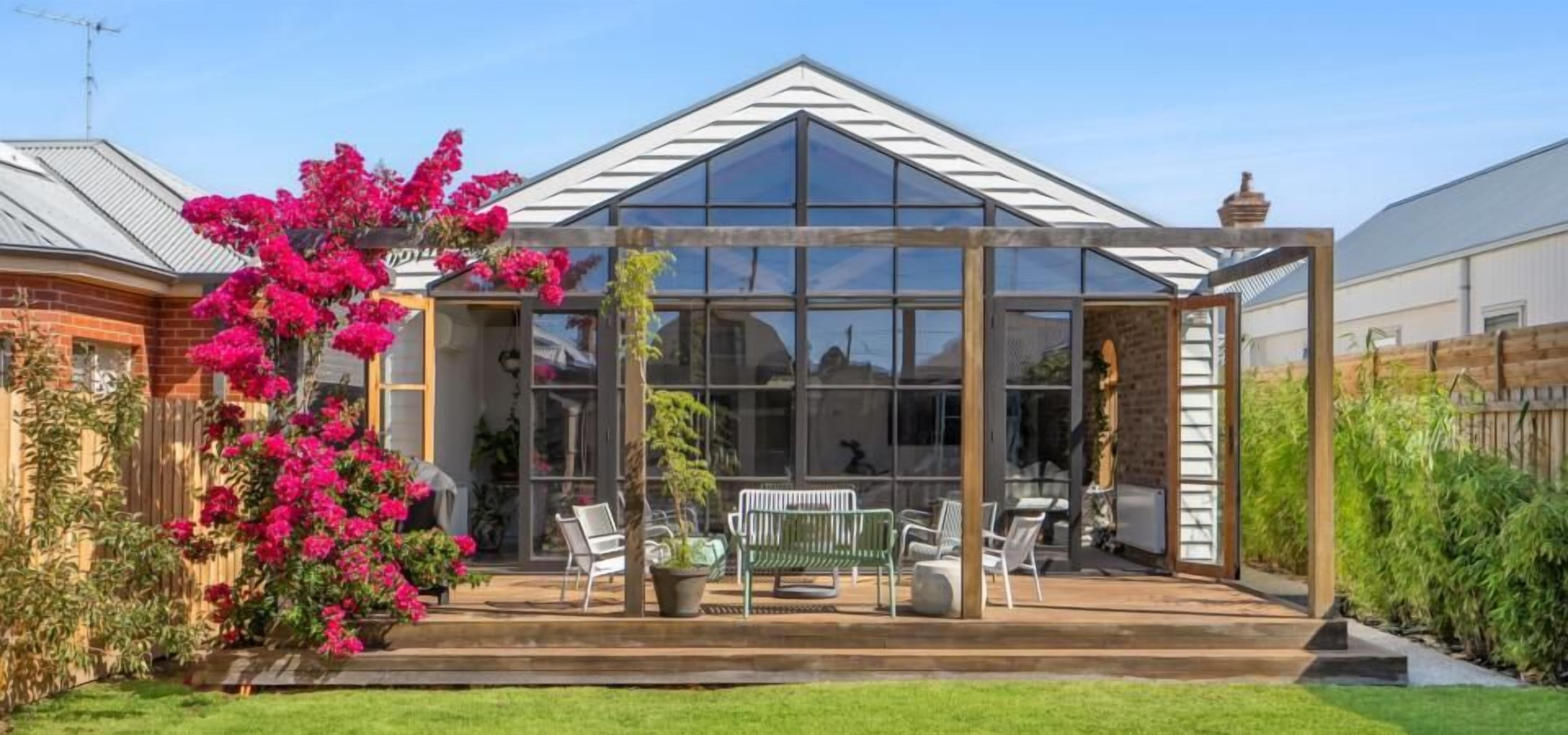 Ein Wintergarten mit Glasdach, umgeben von einer Holzterrasse und einem Garten mit rosa Bougainvillea.