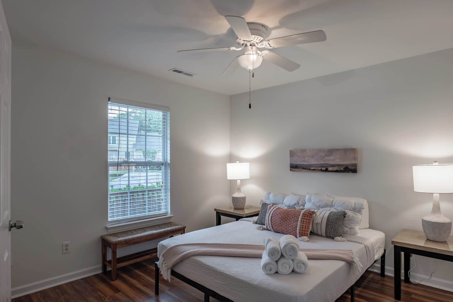 A bedroom with a bed, two bedside tables, a bench, a ceiling fan, a window with blinds, and a painting on the wall.