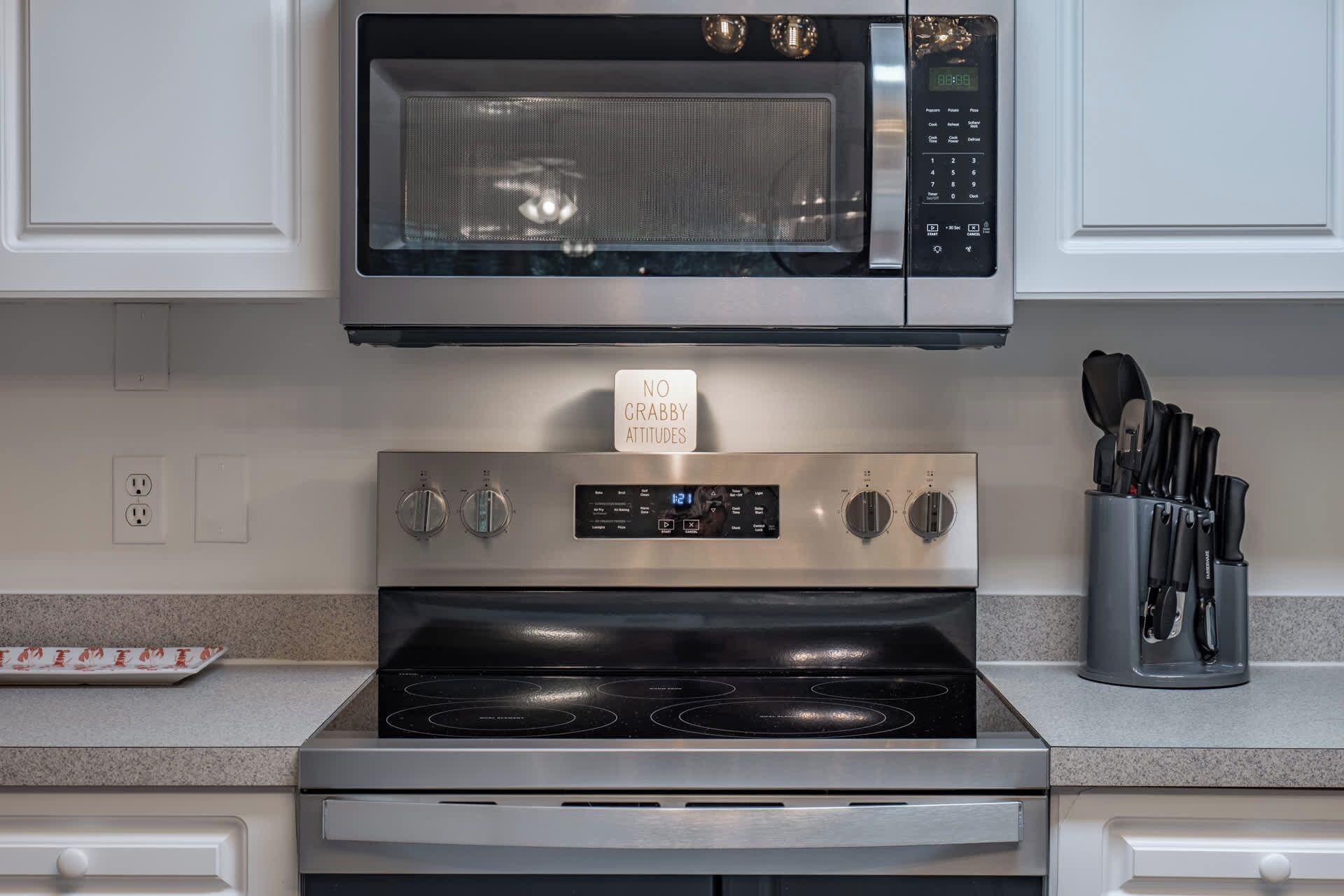 A kitchen with a microwave, stove, and oven. There is a sign that says 'NO GRABBY ATTITUDES' above the stove.