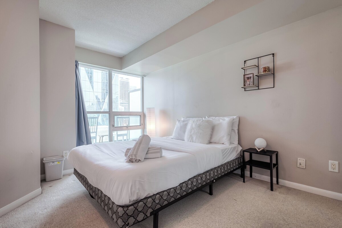 A bedroom with a bed, a nightstand, a wall-mounted shelf, and a window with curtains.