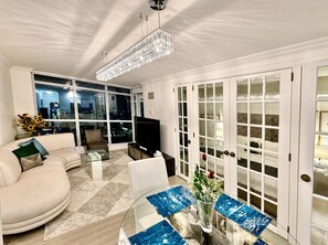 Uma sala de estar moderna com sofá, mesa de centro e televisão. Possui uma grande janela com vista para a cidade e um candelabro.