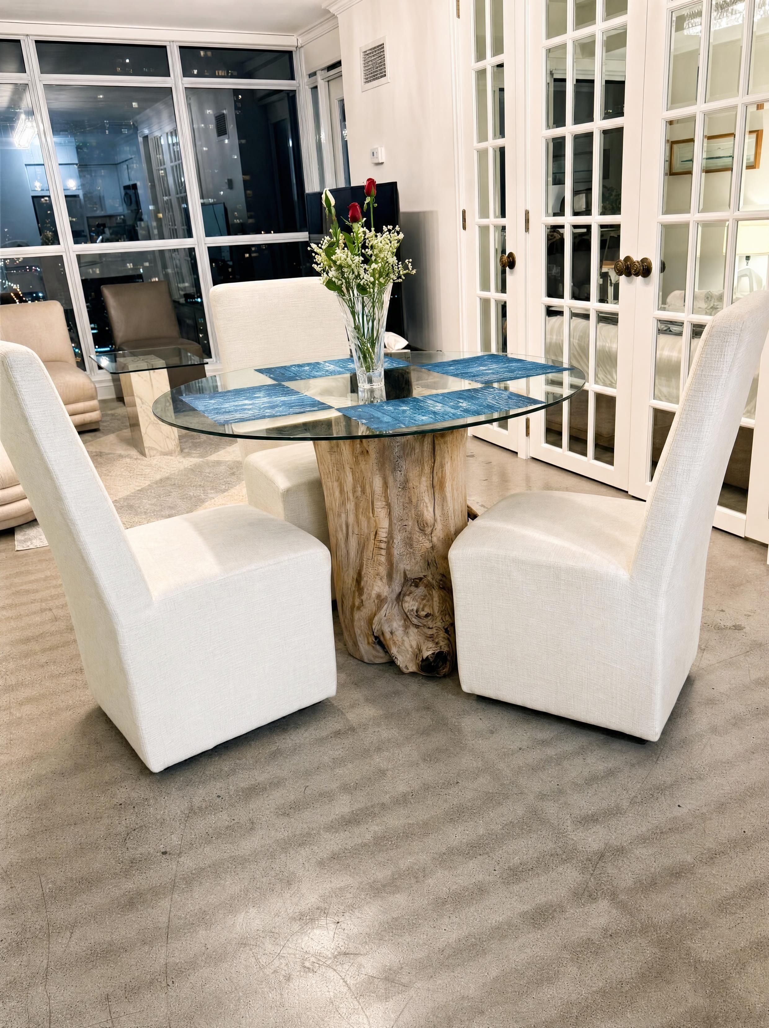 A round glass table with a wooden base, surrounded by two white upholstered chairs.