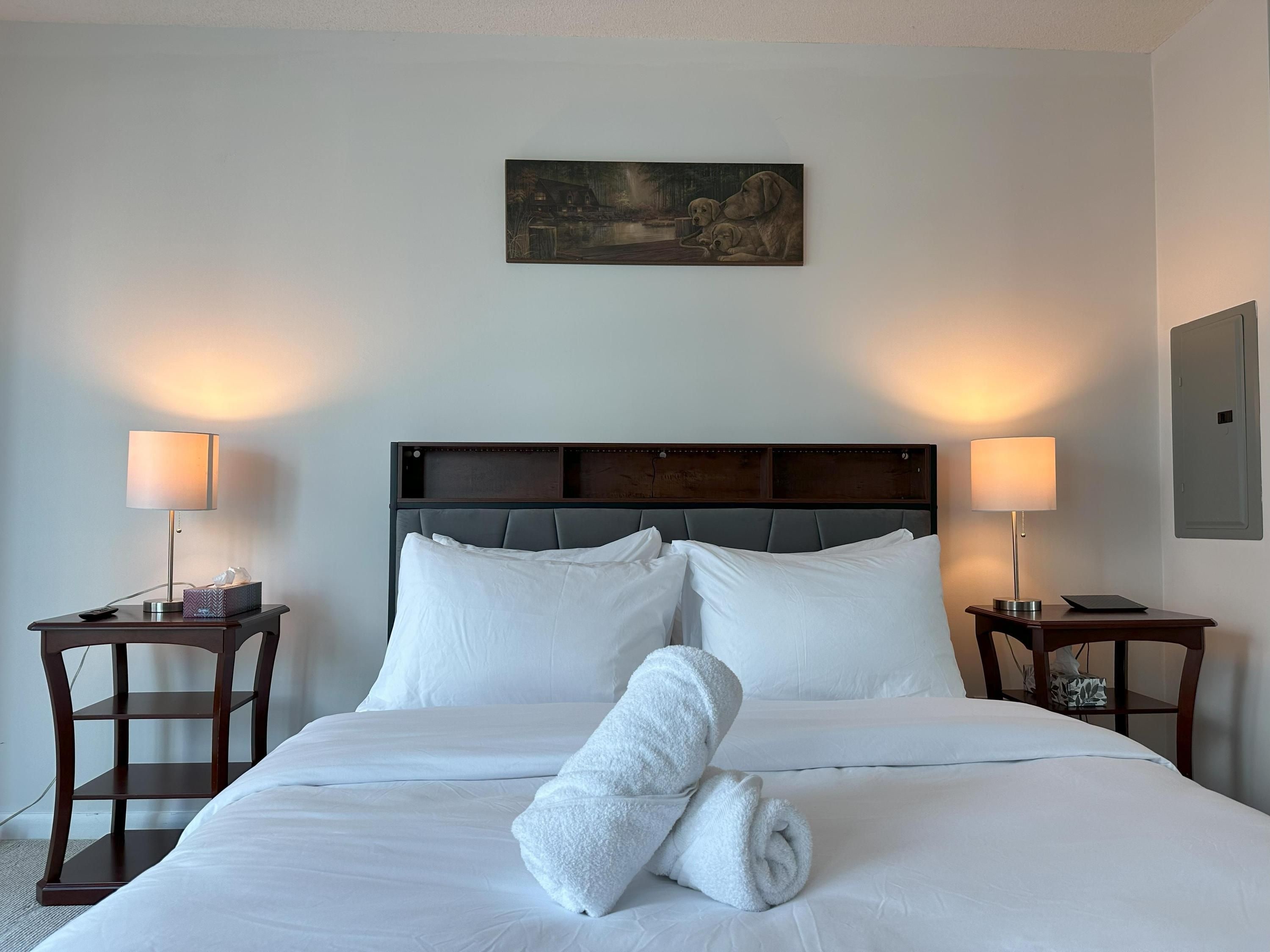 A neatly made bed with white linens, two bedside tables with lamps, and a painting above the bed.