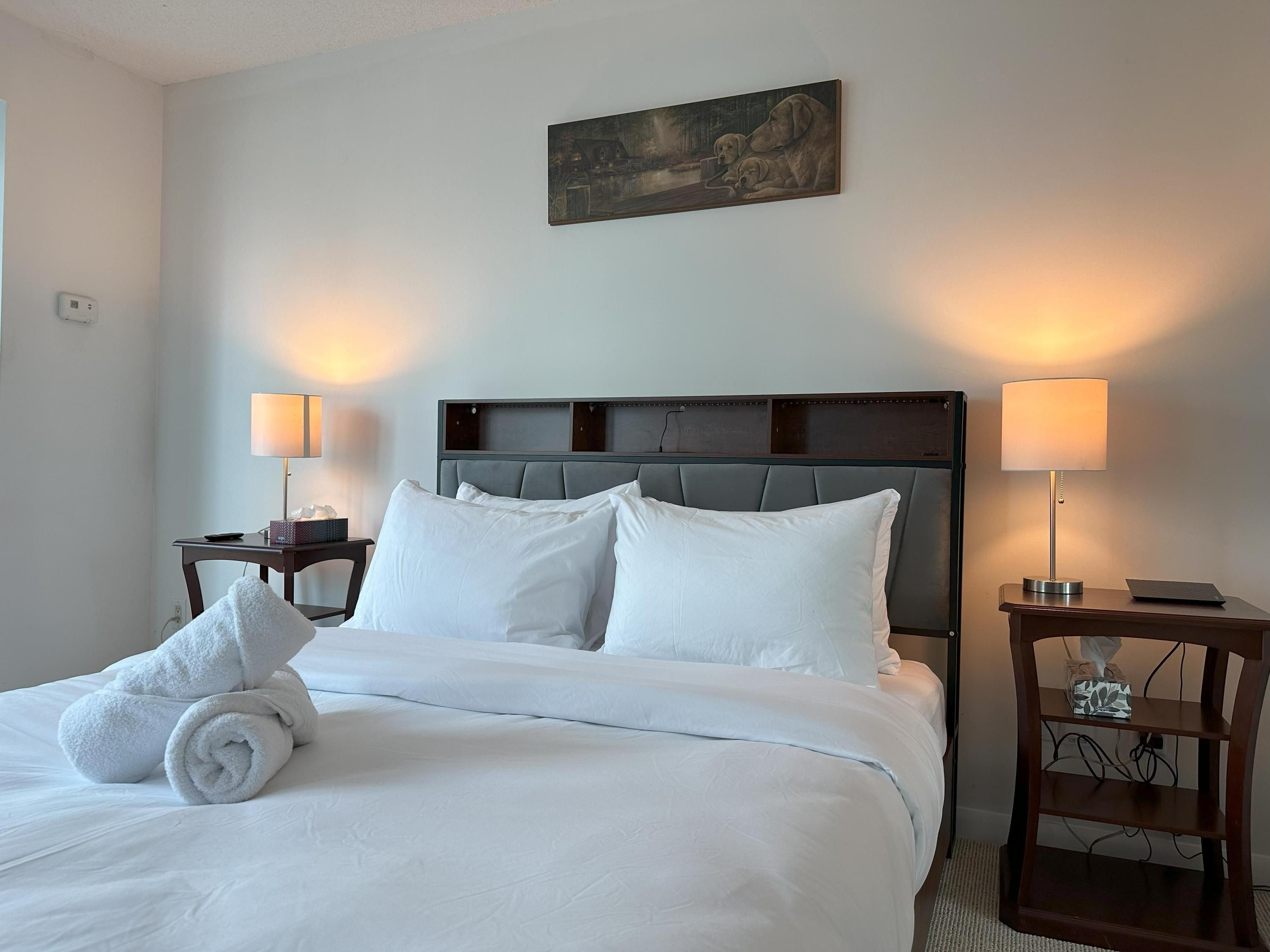 A hotel room with a neatly made bed, two bedside lamps, a painting above the bed, and a wooden nightstand with a tissue box.