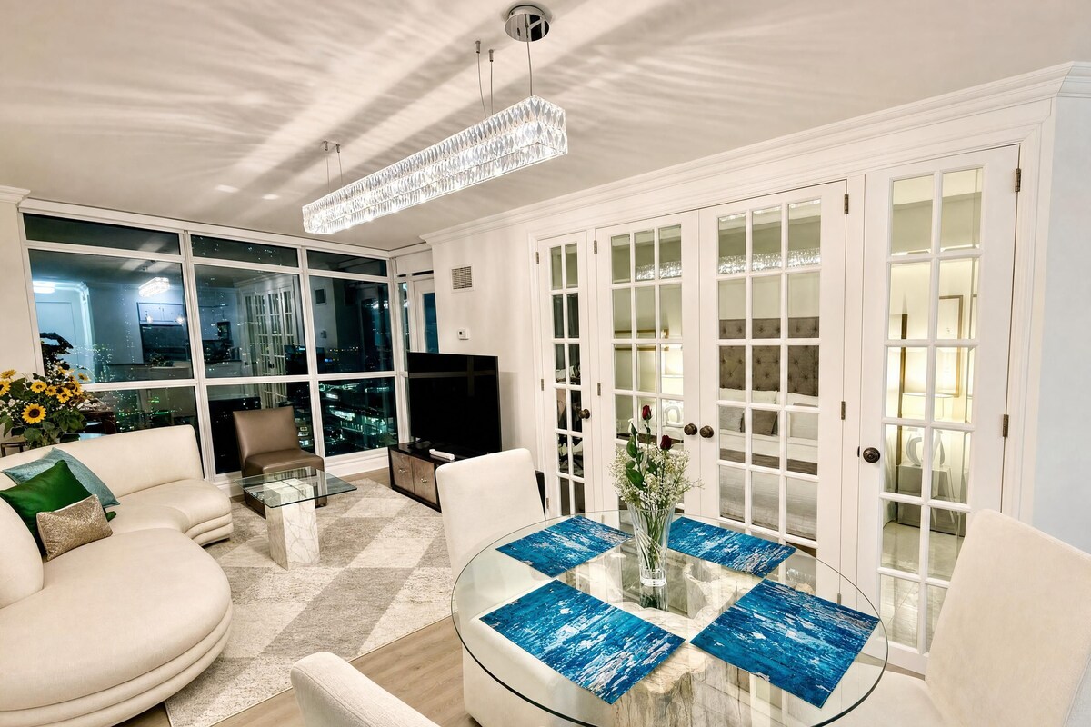 A modern living room with a glass coffee table, white sofas, and a city view through large windows.