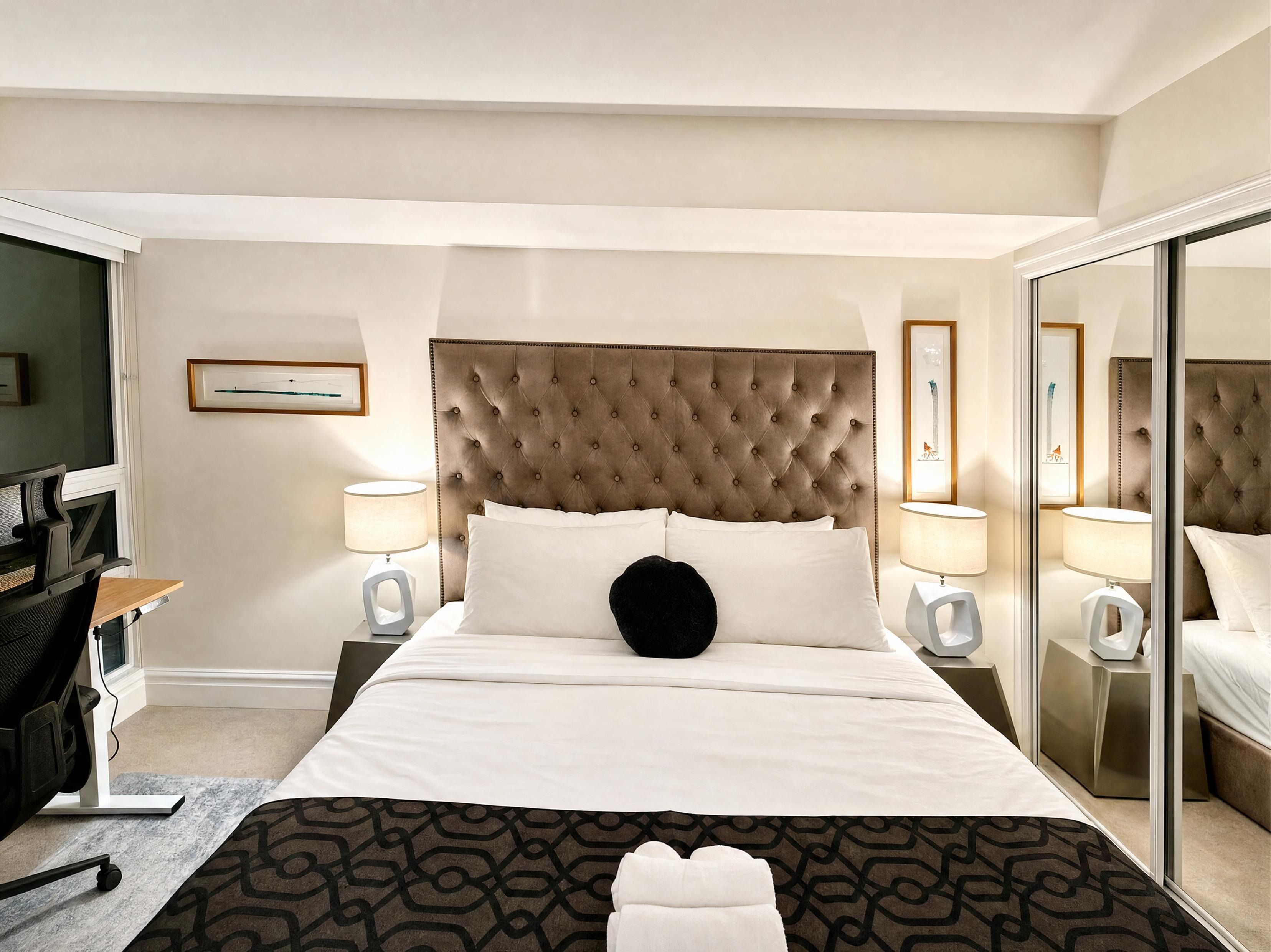 A hotel room with a large bed, two bedside tables with lamps, a desk with a chair, and a large mirror.