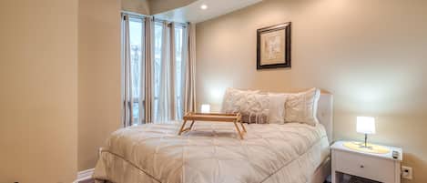 Een slaapkamer met een groot bed, een nachtkastje met een lamp, een lijst met een afbeelding aan de muur en een raam met gordijnen.