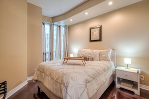 Un dormitorio con una cama grande, una mesita de noche con lámpara, un cuadro enmarcado en la pared y una ventana con cortinas.