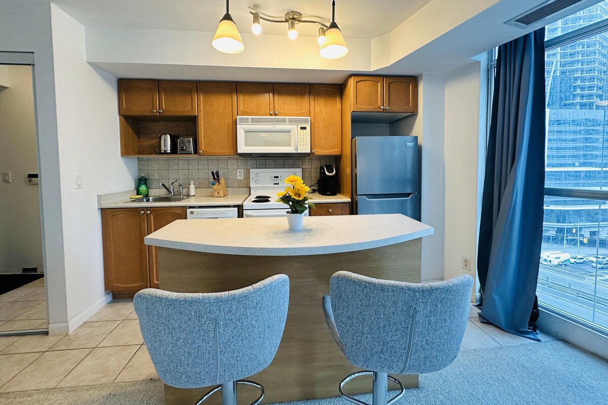 A modern kitchen with wooden cabinets, a white microwave, a stainless steel refrigerator, and a central island with two grey chairs.