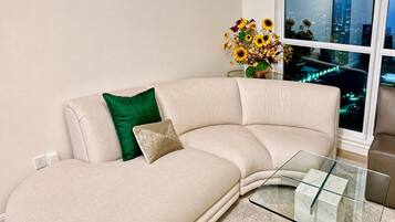 Ein beiges Ecksofa mit einem Glastisch, einem grünen Kissen und einer Vase mit getrockneten Blumen.