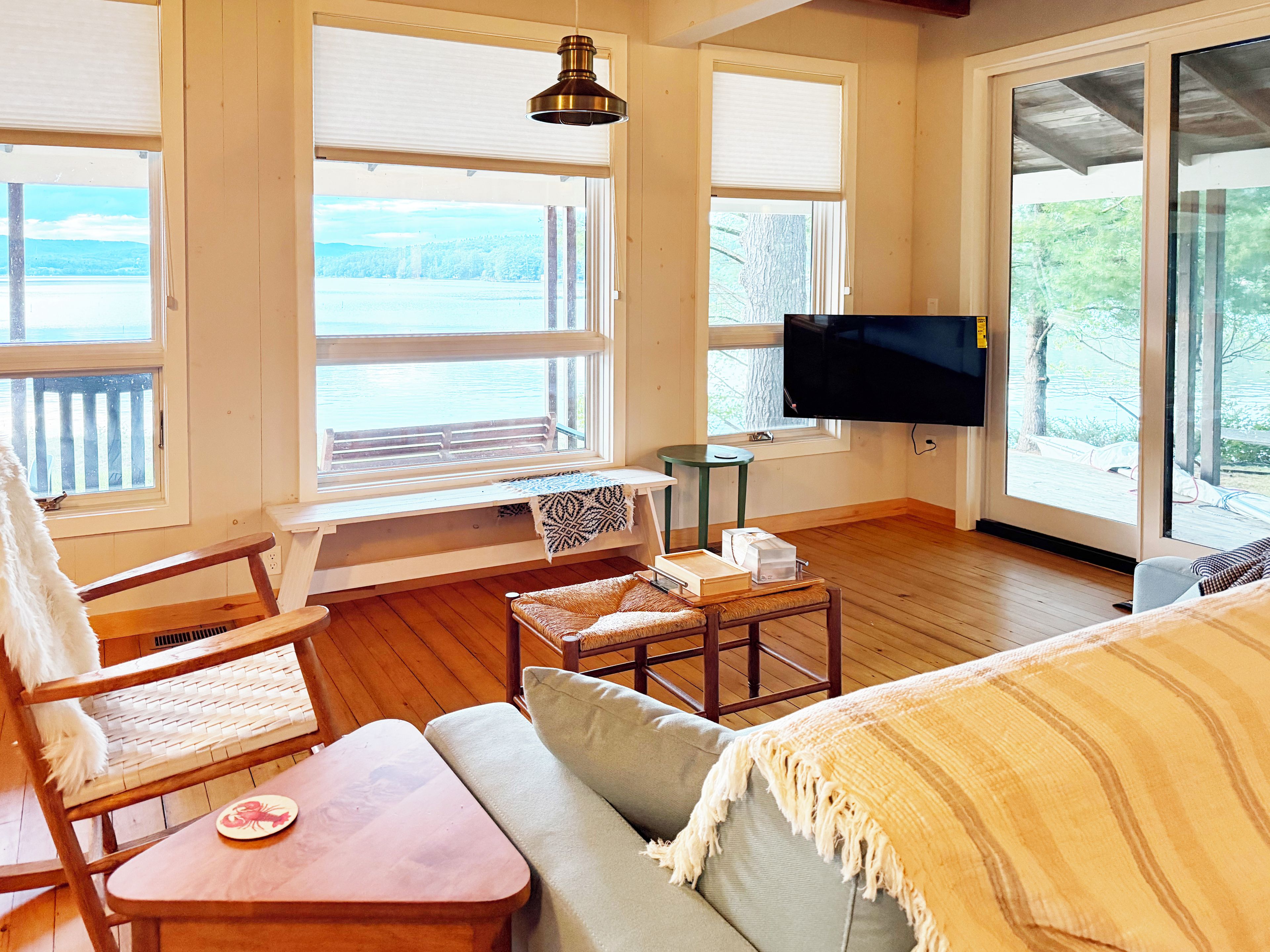 A room with a wooden floor, a sofa, a chair, a small table, a television, and large windows with a view of the outdoors.