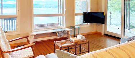 A room with a wooden floor, a sofa, a chair, a small table, a television, and large windows with a view of the outdoors.