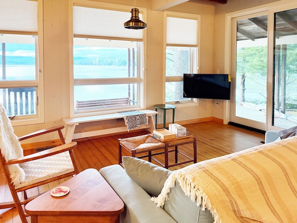 A room with a wooden floor, a sofa, a chair, a small table, a television, and large windows with a view of the outdoors.