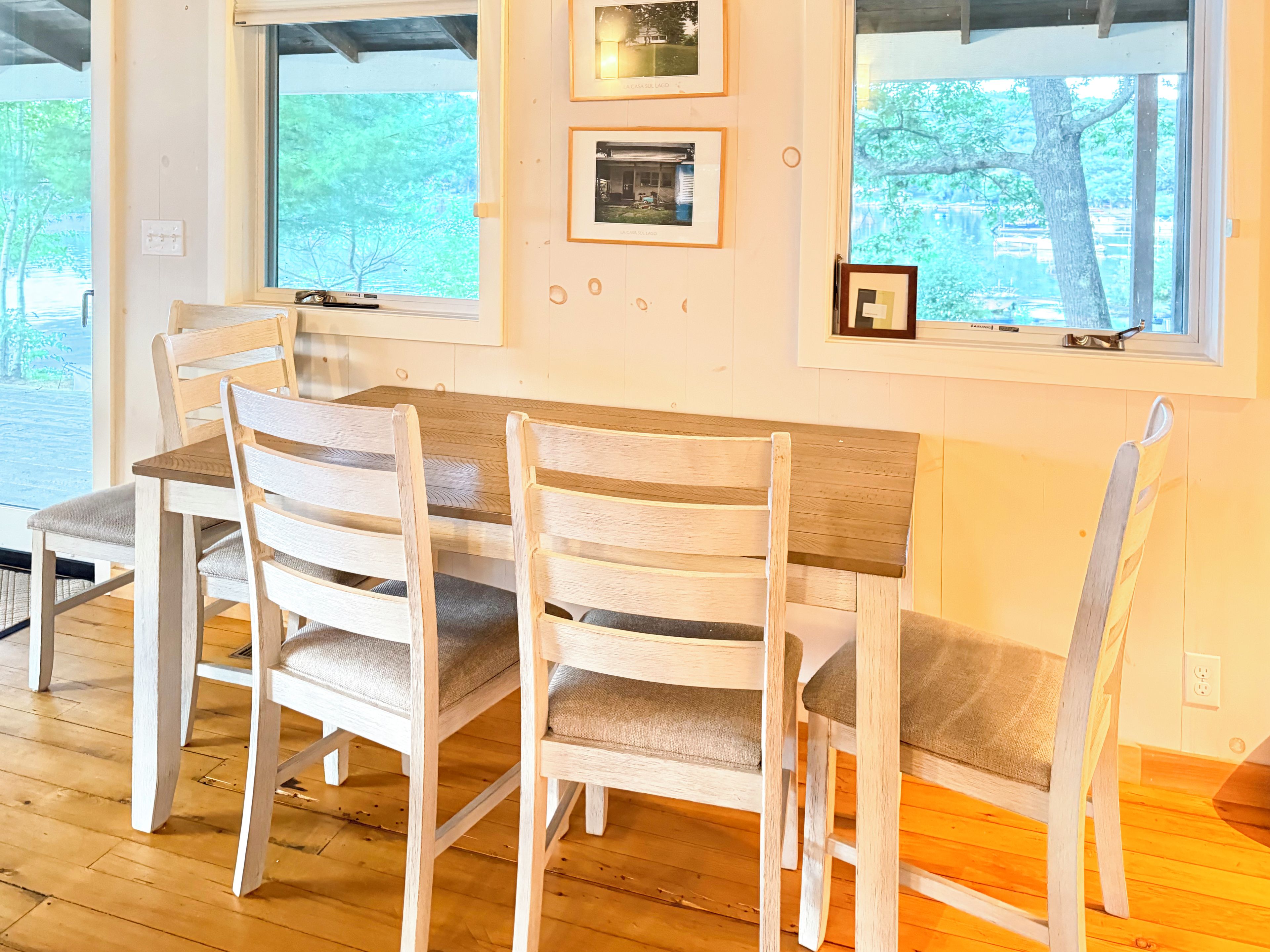 A dining area with a wooden table and chairs, framed pictures on the wall, and large windows.