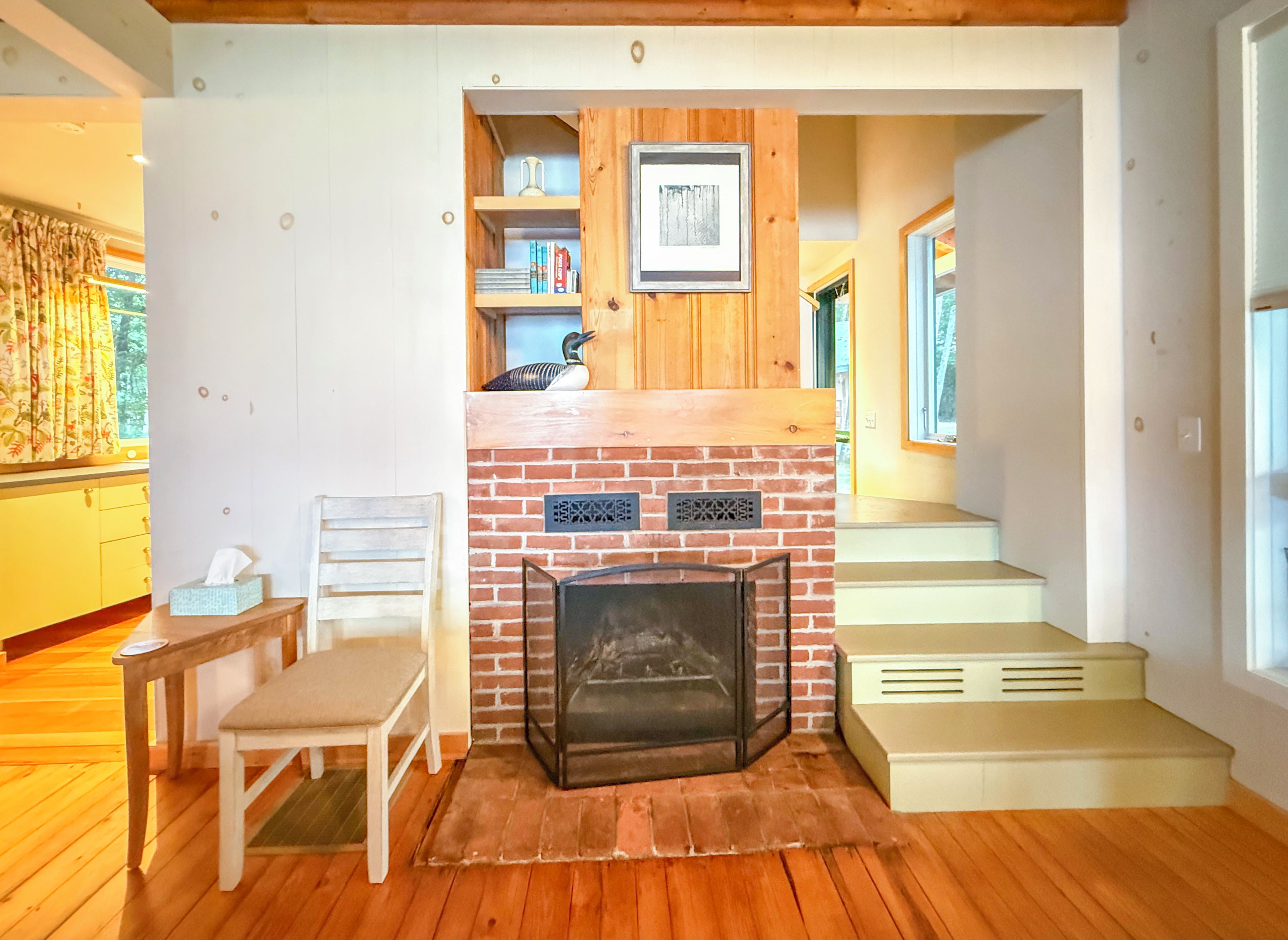 A cozy room with a fireplace, wooden flooring, a chair, a small table, and a staircase leading to an upper level.