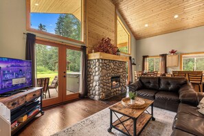 A spacious living room with a stone fireplace, a leather sofa, a television, and a large sliding glass door leading to a deck. - Nature's Acres | Private Hot Tub, Pond & Gazebo (Leavenworth)