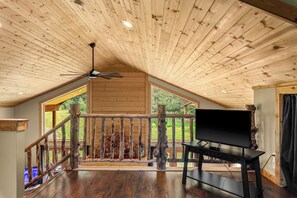 Une pièce spacieuse avec un plafond en bois, un téléviseur posé sur un meuble et un balcon doté d’une balustrade en bois.