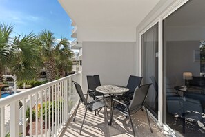 A balcony with a table and chairs, a pool in the background, and a view of a building. - Destin West Gulfside 212 ~ Gulf & Pool Views! (Fort Walton Beach)