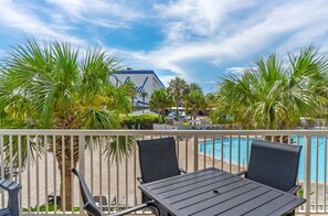 A poolside area with a table and chairs, palm trees, and a building in the background. - Destin West Gulfside 203 ~ Ocean Views ~ Free Beach Service! (Fort Walton Beach)