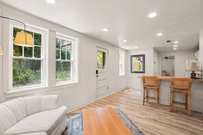 A room with a white sofa, a wooden dining table with two chairs, and a large window with a view of greenery.