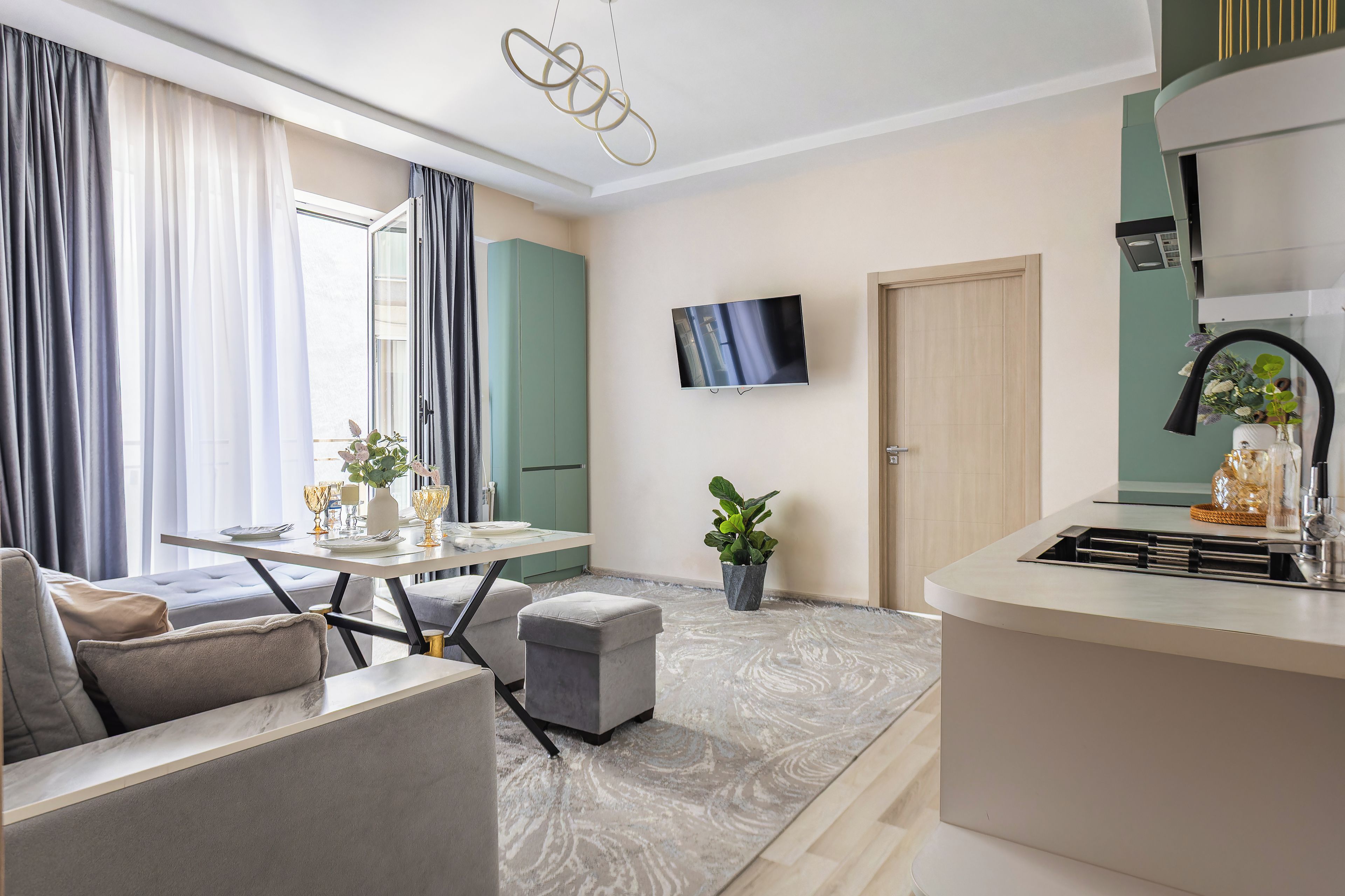 A modern living room with a sofa, a dining table set for two, and a kitchen area with a sink and a flat-screen TV mounted on the wall.