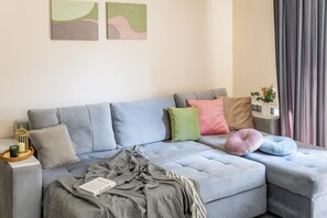 Ein modernes Wohnzimmer mit einem grauen Sofa, mehreren Kissen, einem kleinen Beistelltisch mit Kerze und Blumenvase sowie abstrakten Wandbildern.