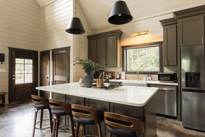 A kitchen with a large island, bar stools, stainless steel appliances, and a view through the window. - Luxe Couples Cabin: Outdoor Cinema+Stargaze Net+Sauna+Plunge+MiniGolf (Broken Bow)