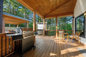 Eine Terrasse im Freien mit einem Grill, einer Sitzgruppe mit Kissen und Blick auf den Wald.