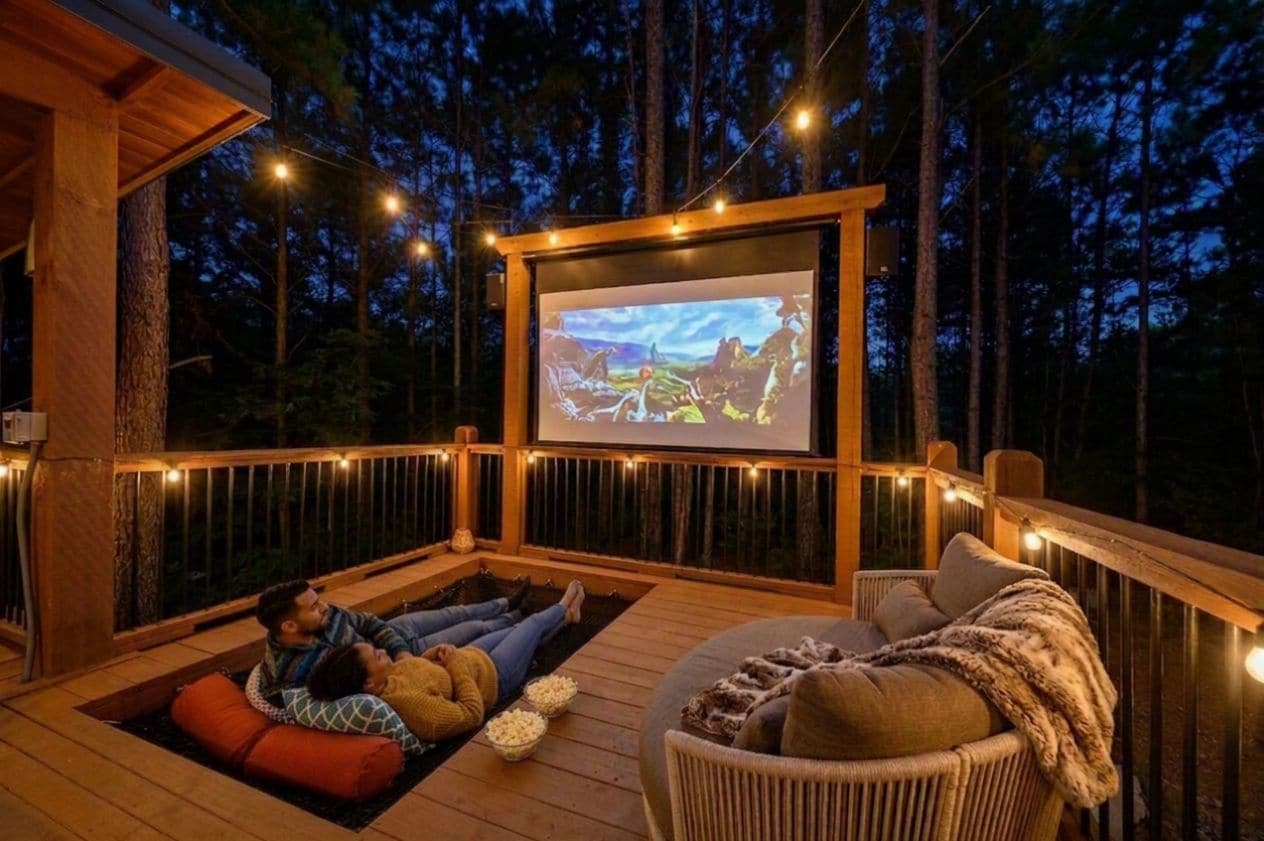 A deck with a screen displaying a movie, surrounded by wooden railings and string lights.