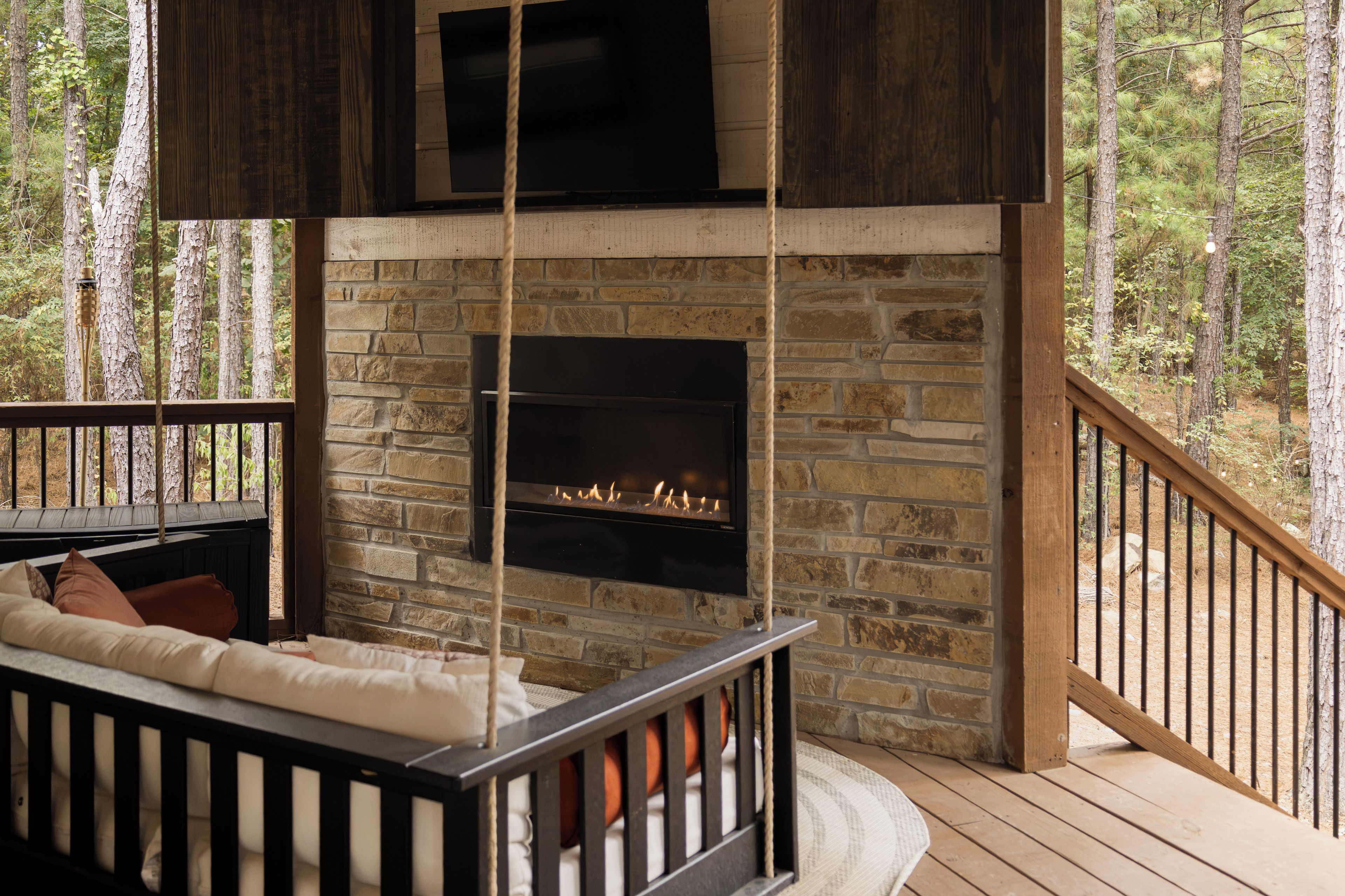 Una chimenea de piedra con un televisor arriba, una terraza de madera y una barandilla con vistas a un bosque.
