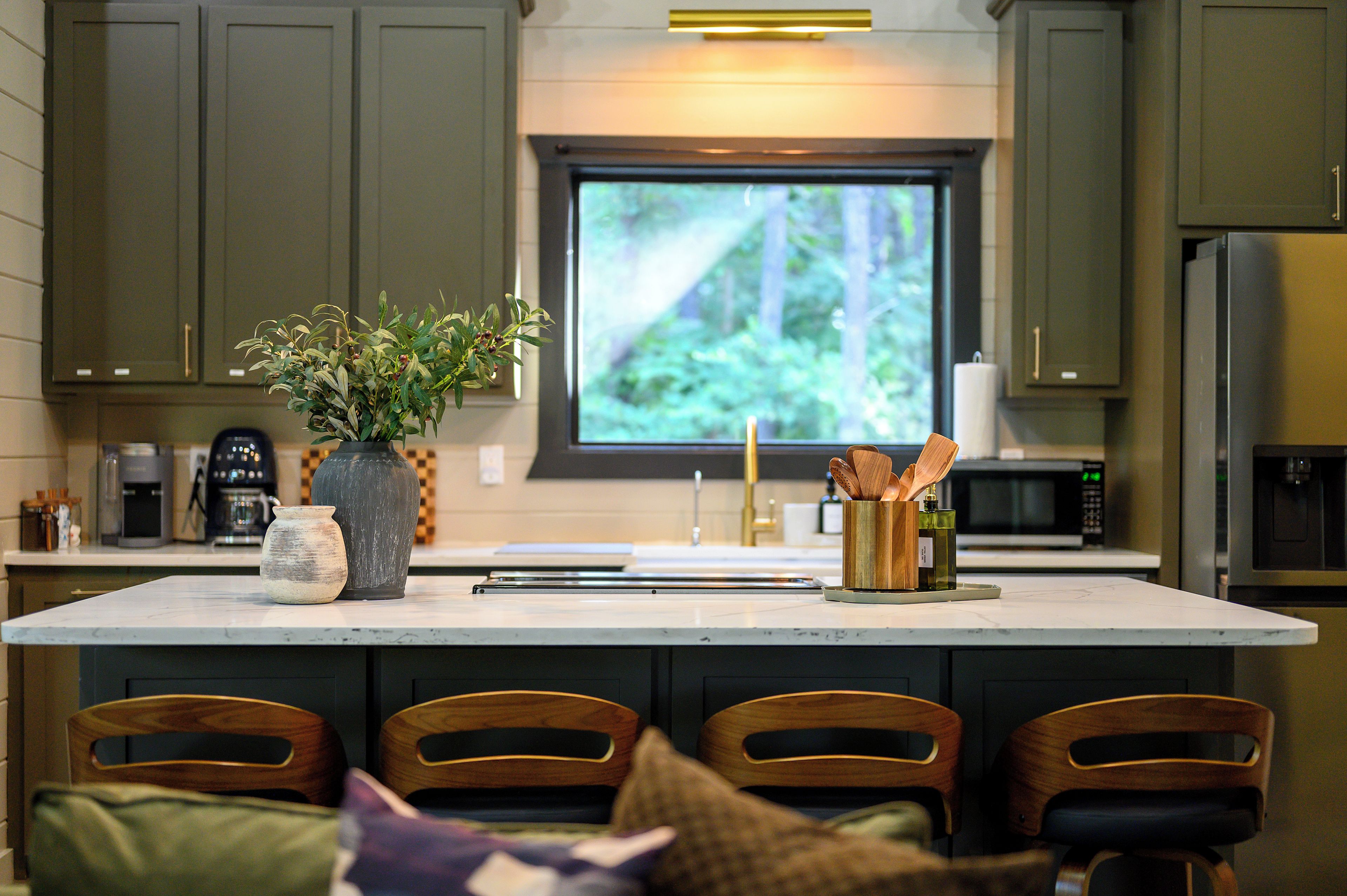 Una cocina con una isla grande, sillas de madera, un florero con follaje y un ventanal con vista al bosque.