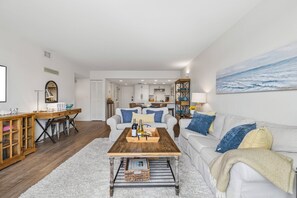 Ein modernes Wohnzimmer mit einem Couchtisch aus Holz, weißen Sofas und einem großen Gemälde, das das Meer zeigt.