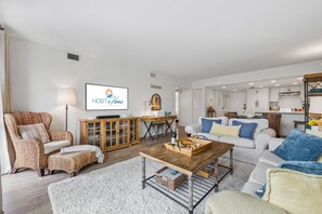 Ein modernes Wohnzimmer mit einem Fernseher, der 'HOST & HOME' zeigt, einem Essbereich mit einem Holztisch und einer Küche im Hintergrund.