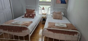 A room with two single beds, each with a metal frame, white bedding, and brown throw blankets. There is a window with blinds, and a small table with a lamp between the beds. - Orontes Cottage Geelong Retreat | Pet Friendly (Greater Geelong)