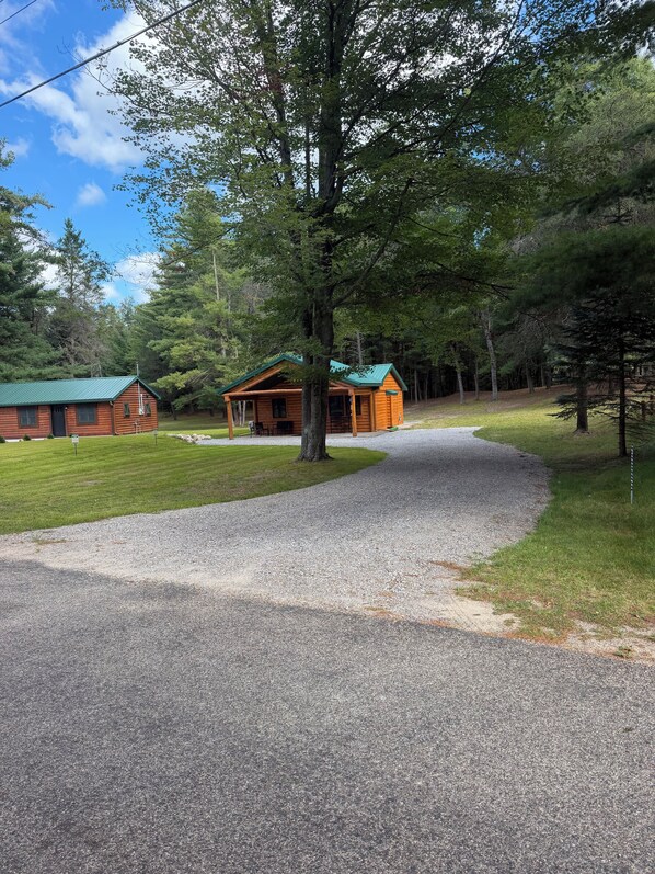 A wooden cabin with a green roof, surrounded by a gravel driveway, and a grassy area with trees.