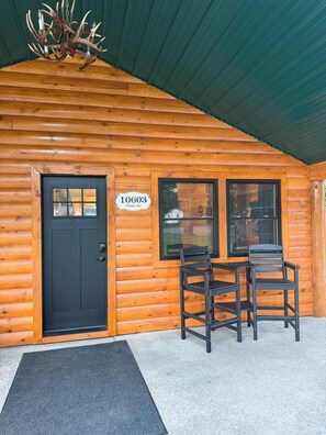A wooden cabin with a black door, a sign reading '10603 Glenn Ave', and two black chairs outside.