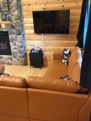 A cozy living room with a stone fireplace, a flat-screen TV mounted on a wooden wall, a leather sofa, and a small black side table with movie tickets.