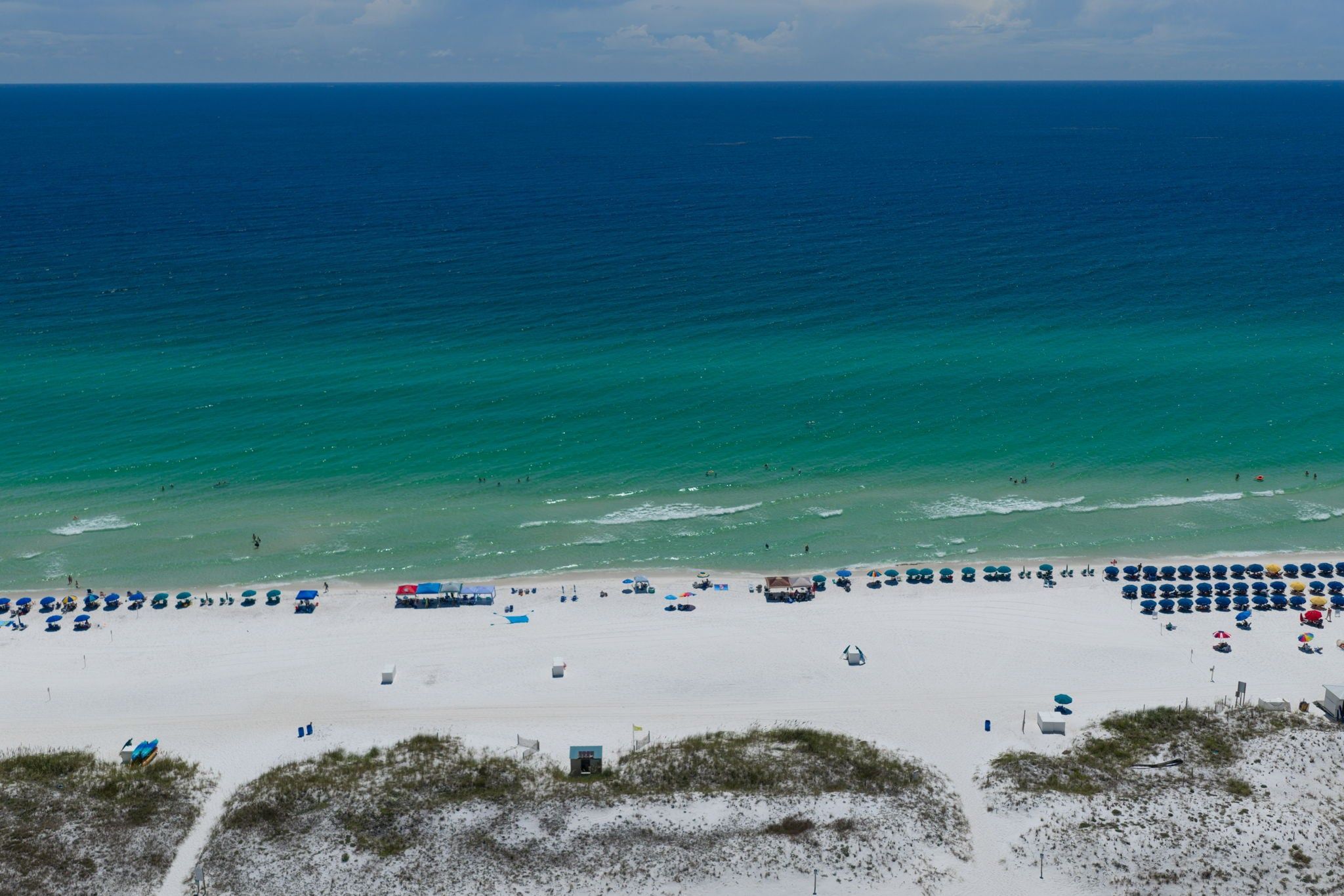 A beach with rows of umbrellas and beach chairs, a sandy shore, and the ocean in the background.