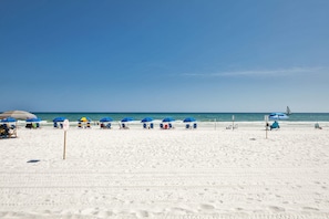 Uma praia com fileiras de guarda-sóis azuis e cadeiras de praia, céu claro e o oceano ao fundo.