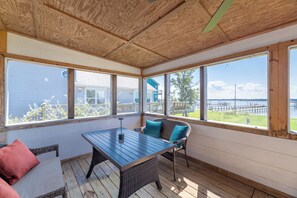 A covered patio with a table and chairs, large windows, and a view of a dock and water. - The Bay House - Soundfront, Dock, Gazebo, Close to Beach, Shops, Restaurants, Flex Stays (Kill Devil Hills)