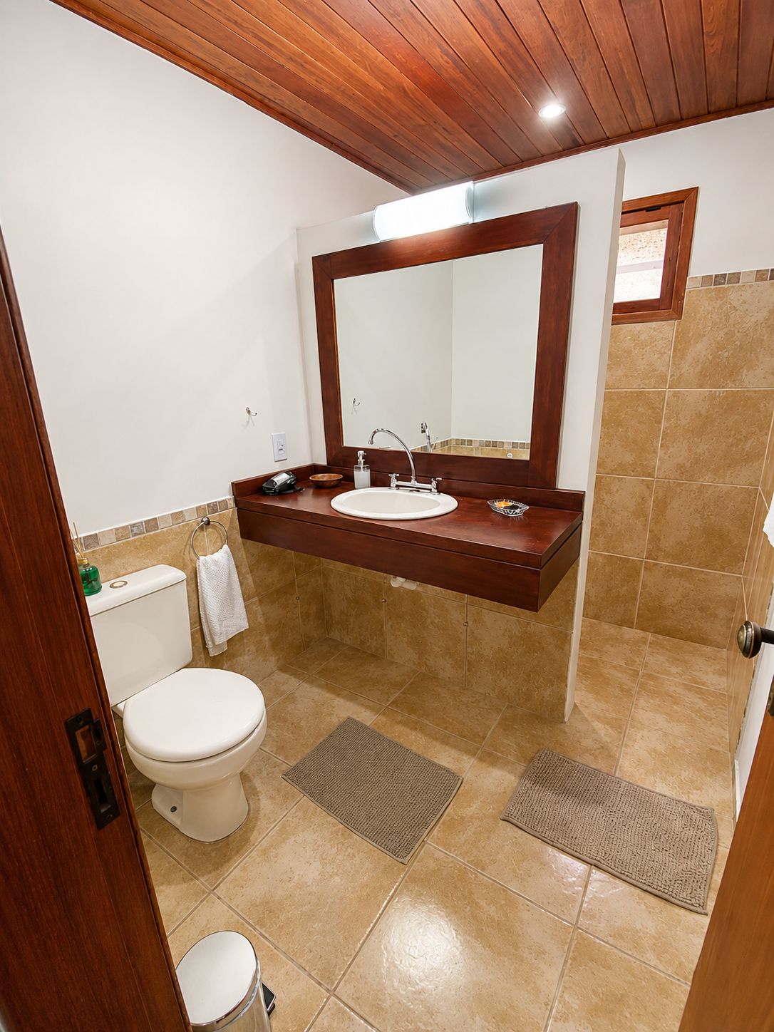 A bathroom with a wooden ceiling, a white toilet, a sink with a mirror above it, and beige tiled flooring.