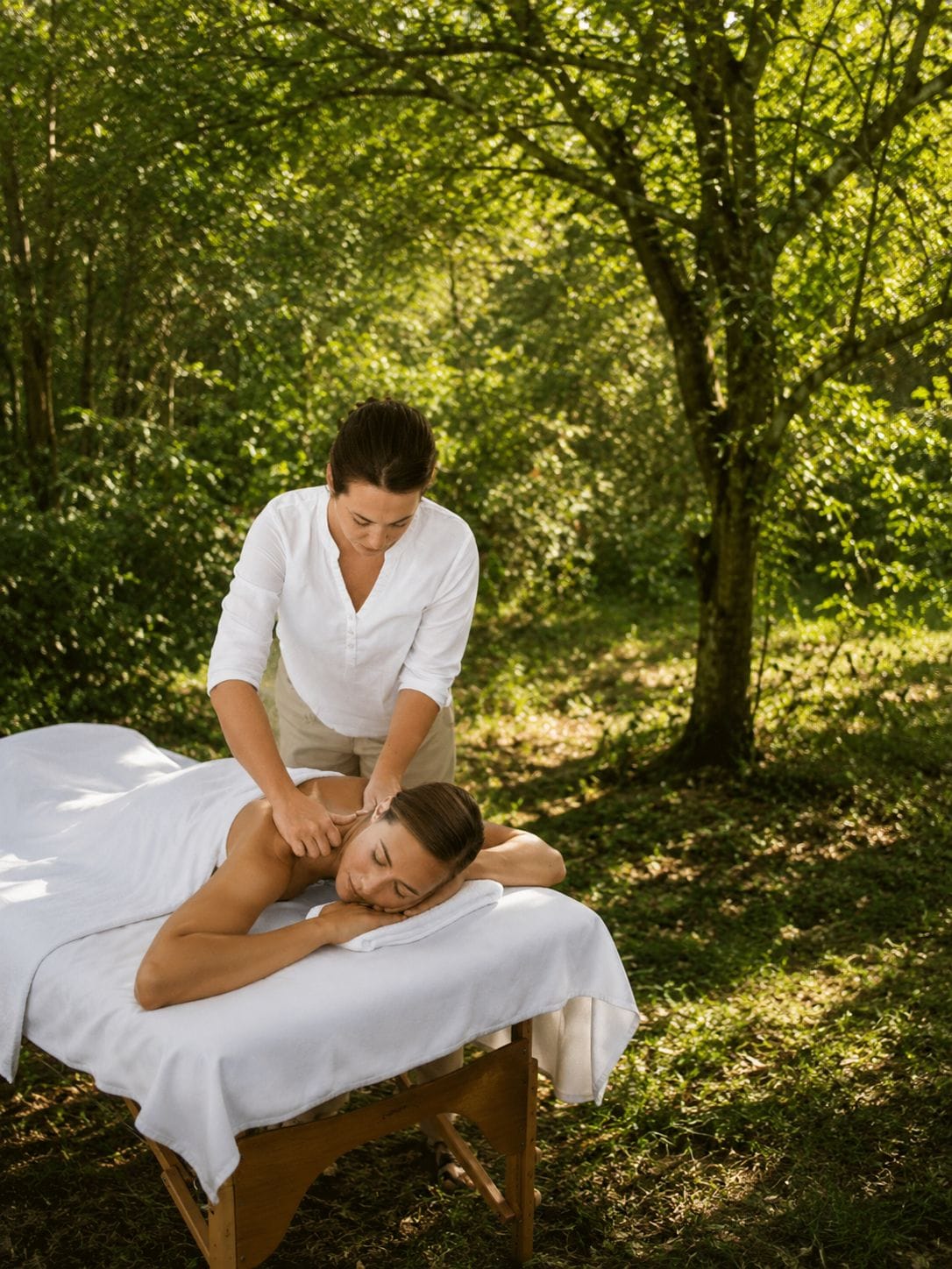 A person receiving a massage outdoors on a wooden platform with a white sheet, surrounded by trees.